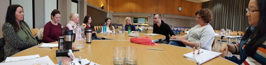 Panoramic shot of people sitting round a long wooden table. There are note books, sheets of paper and coffee cups all across the table.