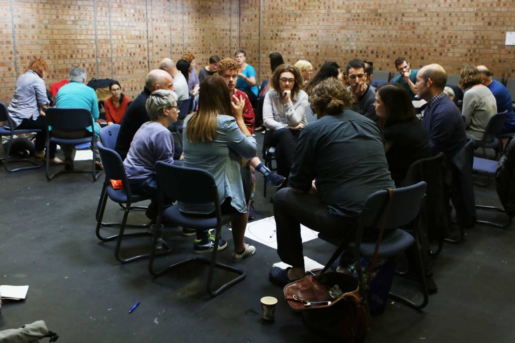 Wide shot of lots of groups of people all sat round in small circles of 6/7 people, each group has a large sheet of paper in the centre of each circle with notes on it.