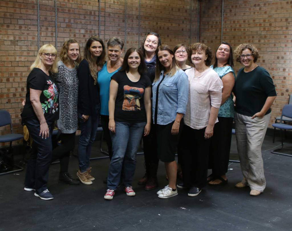 Group shot of 11 women standing in a workshop space, black floor and brick walls. They are all standing, bunched together, smiling at the camera.